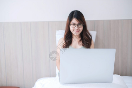 手臂坐着快乐的亚洲女坐在白床上并使用电脑笔记本年轻成女在家工作或网上购物时微笑和欢乐的年轻成女自由者生活方式和技术概念成人亚洲人高清图片素材