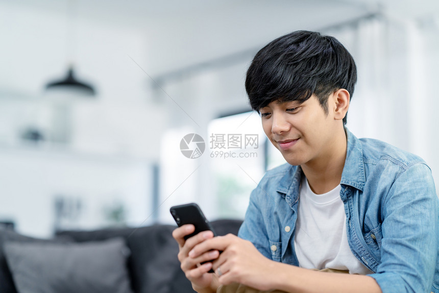 英俊的亚洲人快乐青少年使用智能手机在家中沙发起居室微笑亚洲男子持有并使用手机搜索数据及网上青少年与科技概念的社会中继网Teena图片