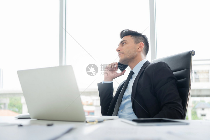 身穿黑西装的聪明帅商正在办公室工作场所使用笔记本电脑工作时坐着用手机与合作伙伴和客户谈判商业往来交易事宜在办公室工作场所从事笔记图片