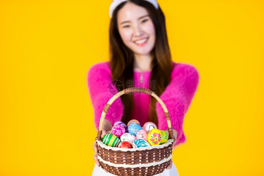 俏皮穿着兔子耳朵的亚洲年轻快乐女在复活节的概念中戴着兔子耳朵的笑容抽象模糊焦点节目则装着一篮子盛彩色复活节鸡蛋在黄色空版间工作室图片