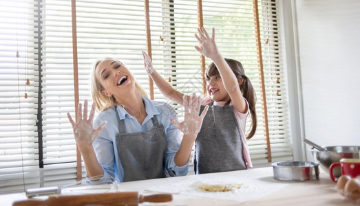 同时提高浓度微笑美丽的年轻母亲和小女儿在厨房的餐桌上笑着做饭玩得开心的家庭同时做蛋糕和饼干快乐的女子和少一起用手边的面粉展示自己幸福生活食物背景