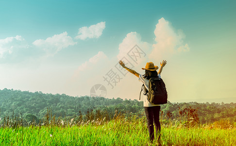 帽子云女爬坡道带着帽子和背包站的快乐女游客在山上举起手来在阳光日照耀的子用绿草地举起手来用蓝天和积雪云彩旅行者享受美丽的风景户外背景