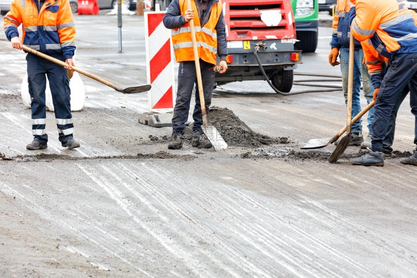 职业工作身穿橙色内裤的公路工人正在用铁铲清洗道路在车上一个有围栏的地区进行清理路工人正在清理车道上有围栏的部分道路街图片