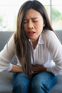 坐在沙发女孩坐在沙发上并忍受腹部因月经泻或消食不良而痛的腹部疼不快亚裔妇女坐落在沙发上忍受着腹部疼痛疾病和保健概念炎恶心康设计图片