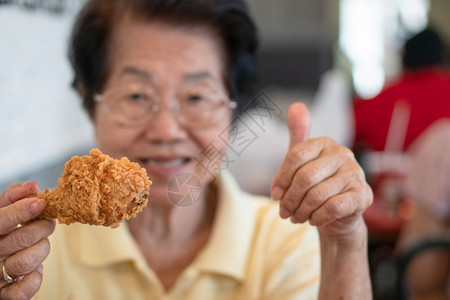 心跳过快有选择地关注炸鸡亚洲老年妇女在餐厅吃炸鸡抬起老龄化社会概念的拇指退休后准备过着幸福的生活笑声在餐馆里亚裔老年妇女正在吃炸鸡享受快背景
