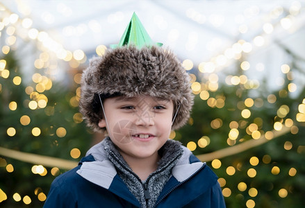 戴花环的男孩新的十二月快乐可爱男孩带着微笑的脸戴党帽面对圣诞树上模糊的灯光Kid在传统圣诞节市场庆祝活动上玩得开心孩子站在明亮的Xmas树旁背景