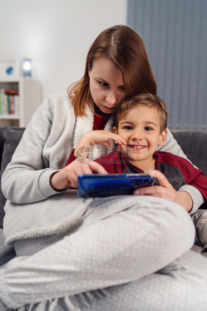 母亲和儿子年轻女母亲和儿子以及她的幼童caucasiansian孩子坐在沙发床上家做视频电话或用手机智能游戏在隔离封闭前视线上播图片