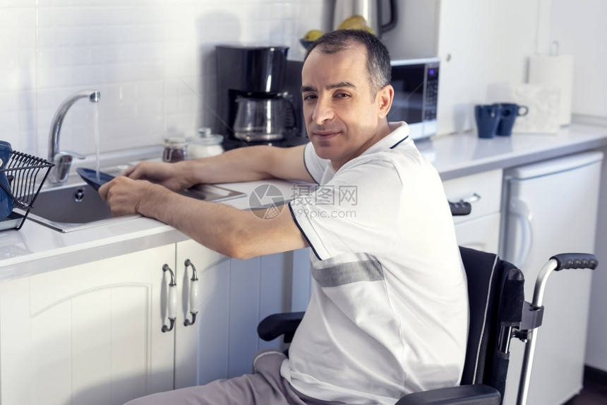 屋重点坐在厨房轮椅上的年轻残疾人在轮椅上洗盘子微笑着年轻残疾人坐在厨房的轮椅上关注他的脸部房间图片