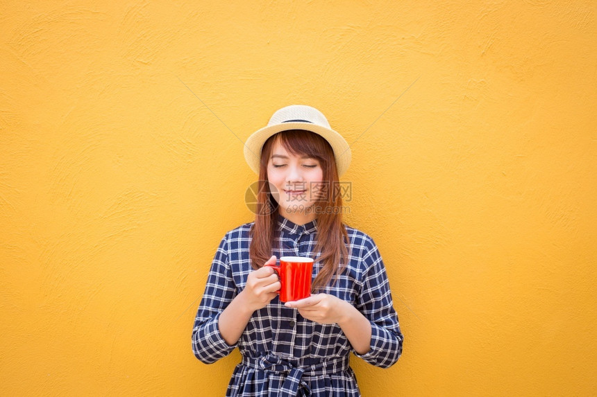 女士保持可爱的带着红色咖啡杯站在黄色的水泥墙背景思想象力上穿着衣服和帽子图片