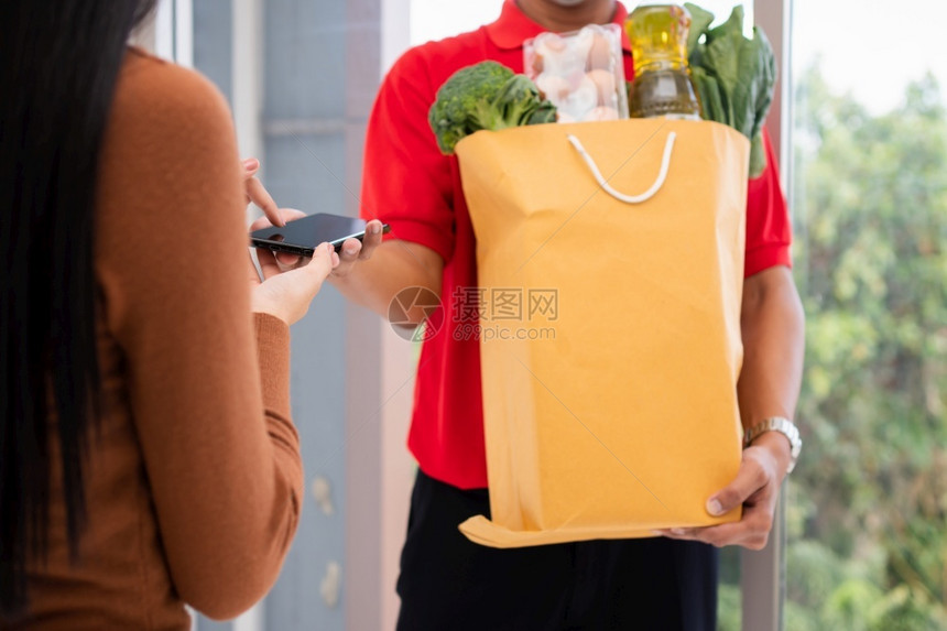 市场距离亚洲送货员持有一袋新鲜食品供顾客使用并拥有智能手机在家中接收付款快速杂货服务和新生活方式概念a提供快餐服务和新生活方式的图片