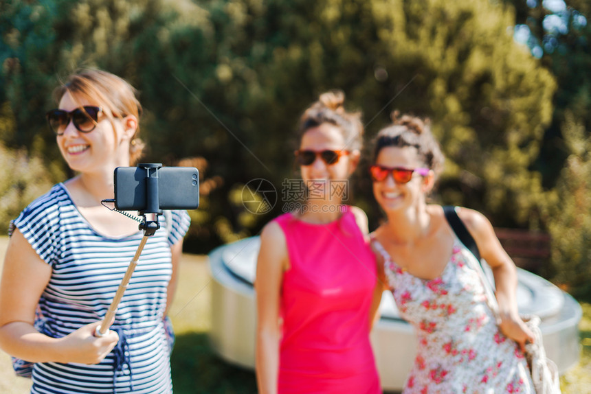 城市的三名年轻女游客使用自拍棍和手机智能在阳光明媚的秋天穿着暑期服装进行自我肖像朋友们照片图片