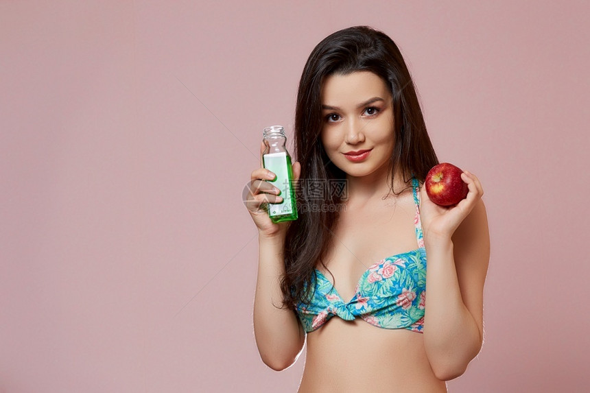 亚洲人身穿泳衣的年轻苗头亚洲女孩带着新鲜果汁和苹在手上微笑粉红孤立的背景上笑幸福健康图片