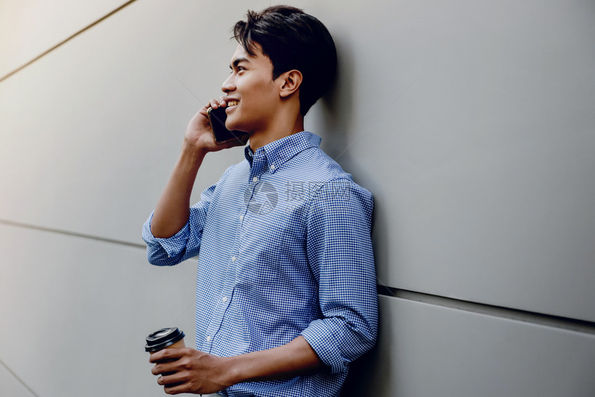 年轻的一位使用现代人移动电话生活方式的快乐青年商人在长墙边站立与咖啡杯的年轻人肖像交流请享用图片