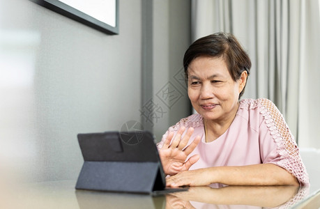 话痨泰国数字的尽管快乐微笑亚洲高龄女在隔绝和家与人隔离时在数字平板电脑上播放视频话穿着粉红色衣服的老年妇女通过互联网技术与人们交谈时背景