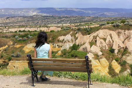 全谷透锥一位身着绿松石色连衣裙的年轻美女坐在山上的木凳观看卡帕多西亚峡谷中山洞和景观的全一位身着绿松石色连衣裙的年轻美女坐在山上的木凳背景