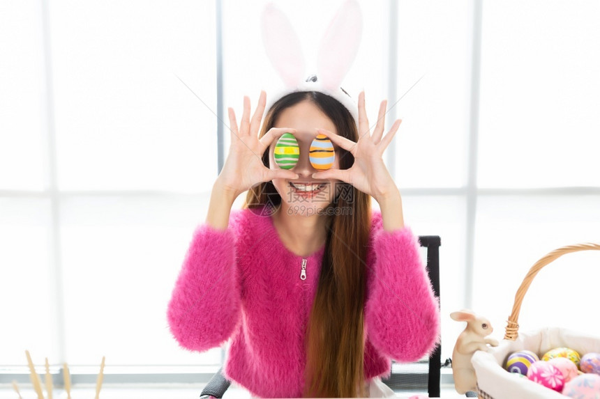 喜悦女士带着兔子耳朵的亚洲年轻女子当着她的眼在白色房间背景中扭曲时在她眼前举起一个多彩的复活节鸡蛋肖像图片