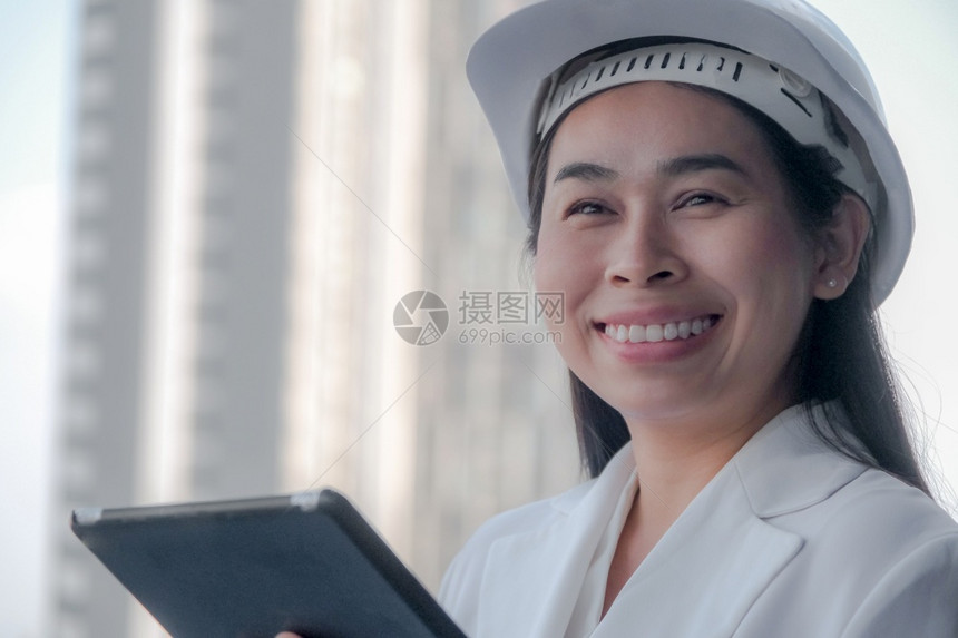 在建筑工地使用平板电脑的女建筑工程师在地用平板电脑对建筑工地进行构和建概念研究时以白头盔向女建筑师微笑自信的技术笔记本电脑图片