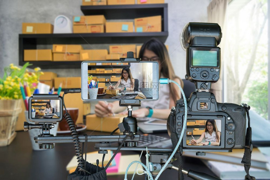 摄像头具有智能移动电话和对年轻亚洲青少业主女商人在家工作的三脚架行动摄像机的专业照相用于网上购物创业者实况流概念为创业者提供Li图片