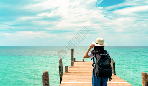 蓝色防伪码美丽的带着草帽和背包放松享受热带天堂海滩的节日女孩站在酒店木制码头上以暑假方式站立在旅馆的木头码上浮桥蓝色的背景