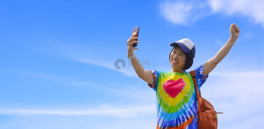 亚洲女旅行者使用智能手机自拍笑与白云和蓝天空背景抗争享受周末假期时举手反对白云和蓝天背景infowhatsthis游客保持背包图片