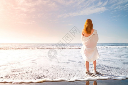 水红头发的嫩女孩赤脚站在海滩上的白色冲浪泡沫中穿着白色连衣裙的女人在海边阳光明媚的早晨红头发嫩女孩赤脚站在海滩上的白色冲浪泡沫中背景图片