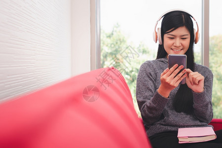 白种人房间技术清晨坐在沙发上的亚洲可爱女人坐在沙发上用手机送信息同时听音乐在客厅大腿上放一本书笑声背景图片