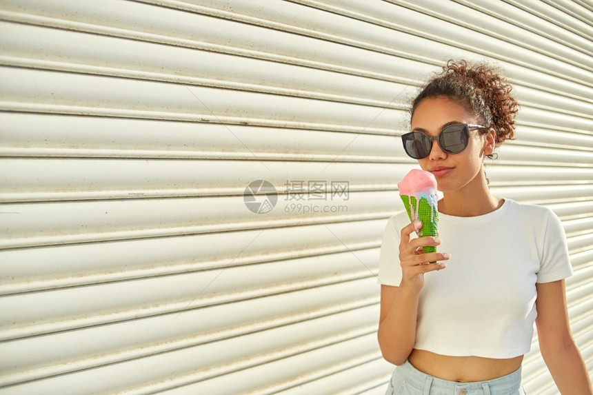 幸福天奶油穿着白色T恤和轻牛仔裤的年美籍黑人女孩在日光照耀时用冰淇淋抵挡着一堵轻墙吃冰淇淋图片