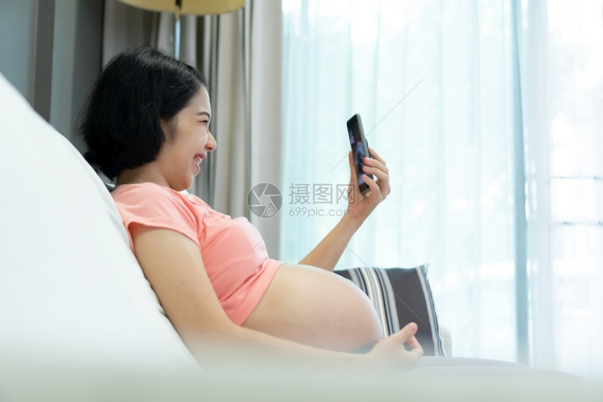 孩子有吸引力的亚洲孕妇穿粉红色衬衫在线视频电话与家庭交谈时使用手机在怀孕互联网和通讯中用智能手机聊天快乐微笑的年轻母亲容Happ图片