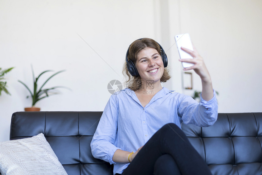 快乐的坐在沙发上做自拍的年轻美女在她的手机上坐着摄影图片