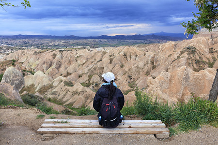 砂岩一个年轻女孩背着一个旅游包坐在木板凳上看着山峡谷和暴风雪一个年轻女孩坐在木板凳上看着地平线卡帕多西亚山脉和风暴天空亚洲木制的背景图片