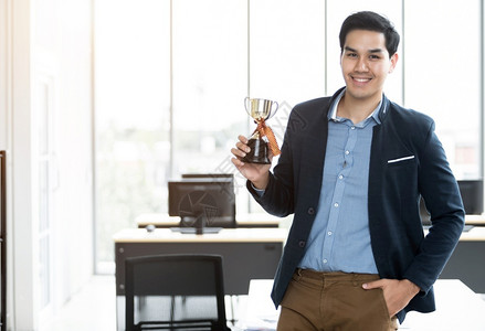 自信的最好幸福成功感觉胜利者肖像尖叫着英俊的年轻亚洲商人在办公室背景中拿着冠军杯在办公室背景背景图片
