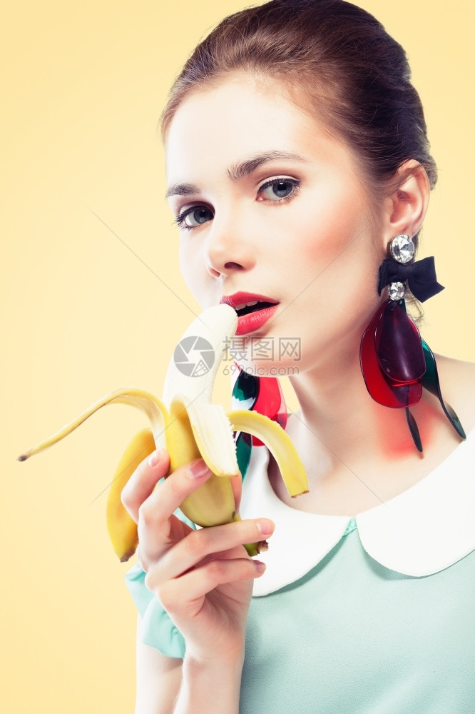年轻美女嘴唇和眼箭红色的美容粉辣妹化妆时戴漂亮的塑料耳环吃香蕉黄色背景针纹风格拉普捷夫感的白种人图片