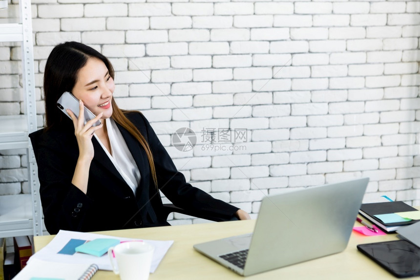 成功快乐的亚裔年轻女商人抓住手持智能机用笔记本电脑在白色木桌墙办公室背景的白木桌子上工作学生吸引人的工作场所图片