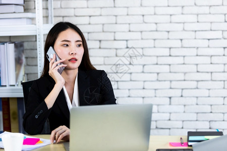 细胞成功快乐的亚裔年轻女商人抓住手持智能机用笔记本电脑在白色木桌墙办公室背景的白木桌子上工作女孩场所背景图片