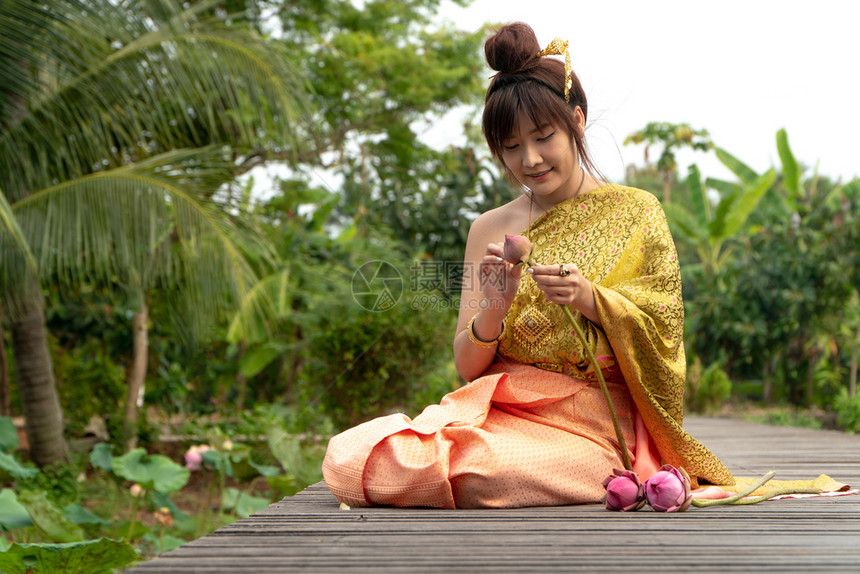 拥有绿色身着传统泰式礼服坐在木桥上的女美丽她手在泰国风格下举手受人尊敬在莲花叶上还有粉红莲亚洲图片