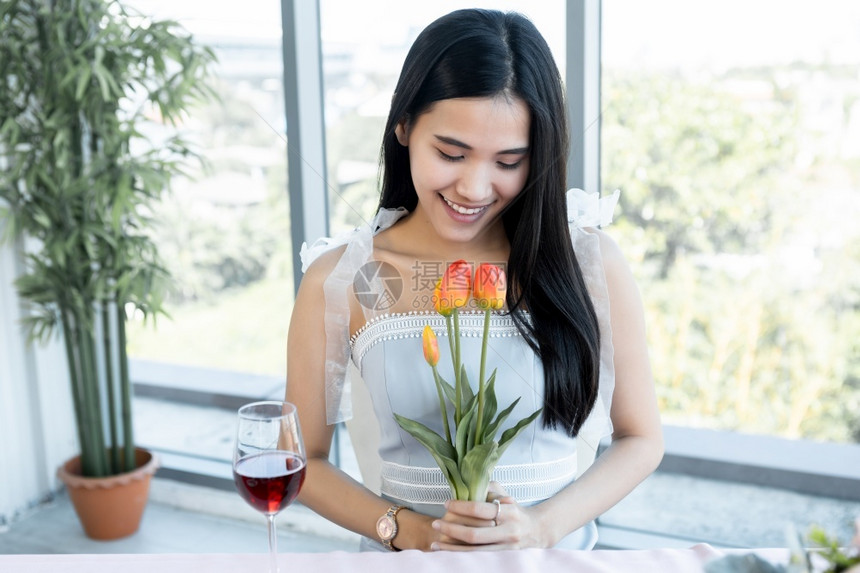 成人开心地笑着的亚裔年轻女坐在餐桌上吃饭厅背景中拿着一束郁金香花亚洲人颜色图片