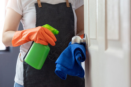 胚芽家庭女佣用橙色橡皮手套在门的触摸面上打扫门把手用蓝色微纤维清洁布和绿色防菌喷雾瓶用于卫生的CoronaCOVID19预防概念背景图片