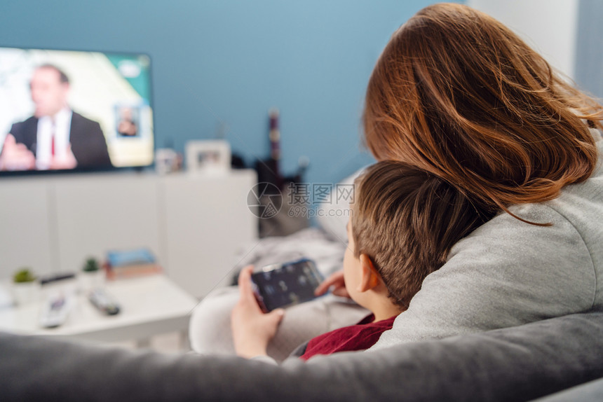 在室内母亲和儿子年轻女母亲和儿子以及她的男孩Caucasiansian孩子坐在沙发床上家做视频电话或玩子游戏在手机智能上播放视频图片