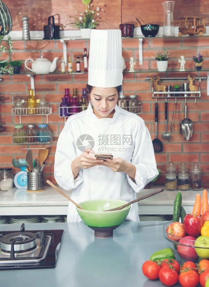 年轻女厨师黑头发穿白西装色制服看厨房的智能手机消息传递当代的空图片