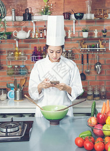 年轻女厨师黑头发穿白西装色制服看厨房的智能手机消息传递当代的空图片