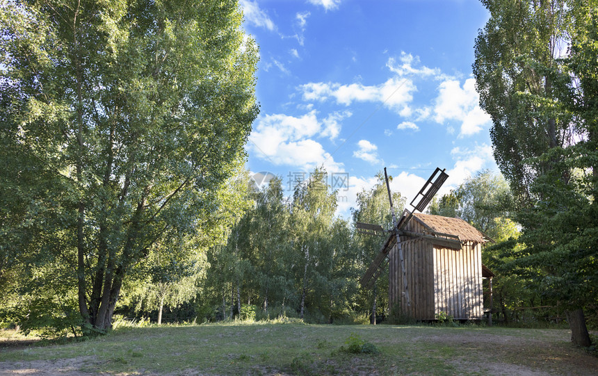 一种座古老的木制风车在青春的森林里一个绿色的草地上面对着一片蓝色的天空和浅白云一座古老的木制风车在夏日的森林里生长农场图片