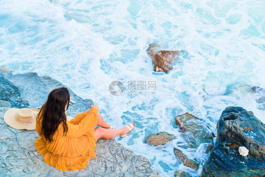 快乐的假期日落时年轻美女后退时看晚上穿着衣服的女孩在海边日落背景的岩石上年轻美丽的女孩魅力图片