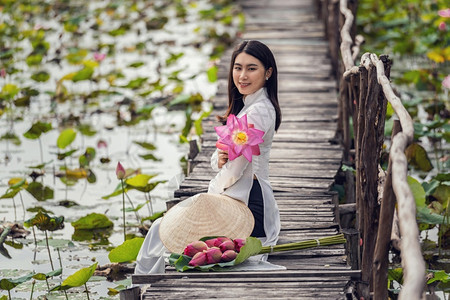 桥上女孩在大莲花湖维特南青山或东方的木桥上挂着粉色莲花的传统维特南帽子坐在木桥上的美丽女肖像绿色坐着敖背景