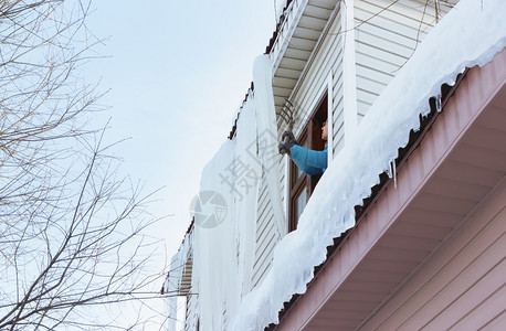 屋顶雪工作一个人从屋顶上搬走巨型冰柱和雪从窗户穿过在农村的季节建筑维护概念中使用干草叉在明冬日用洞叉把大冰柱和雪从窗边移走白色的建造背景