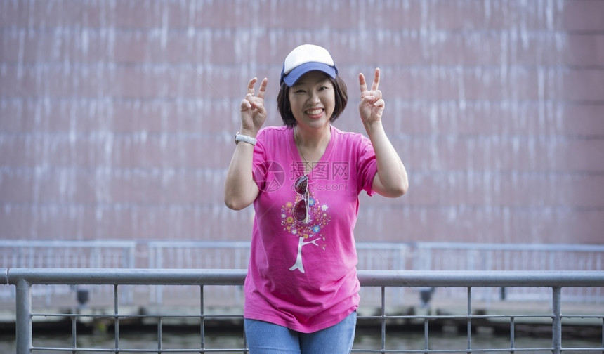 愉快栏杆脸亚洲乐的女游客展示两只胜利手的标志同时微笑着在公共园建筑墙上的瀑布装饰前拍摄影机图片