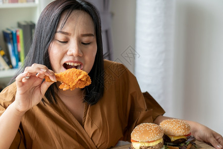 百吃不厌包子小吃饥饿的超重女持有炸鸡木板上的汉堡包和桌披萨在家工作期间体重增加问题百食不厌症BED概念成人背景