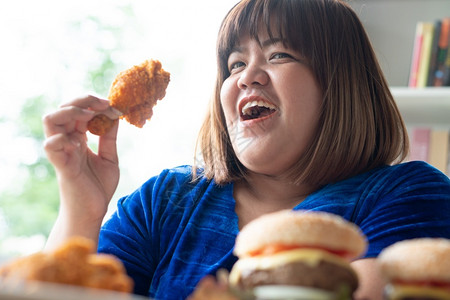 盘子加大尺码一顿饭饥饿的超重女持有炸鸡木板上的汉堡包和桌披萨在家工作期间体重增加问题百食不厌症BED概念背景图片