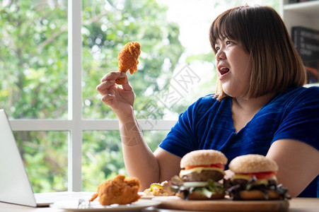 百吃不厌垃圾美味的饥饿超重女持有炸鸡木板上的汉堡包和桌披萨在家工作期间体重增加问题百食不厌症BED概念健康背景