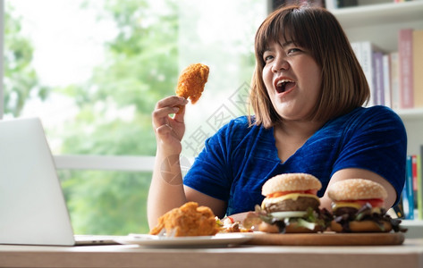 肥胖手午餐饥饿的超重女持有炸鸡木板上的汉堡包和桌披萨在家工作期间体重增加问题百食不厌症BED概念背景图片