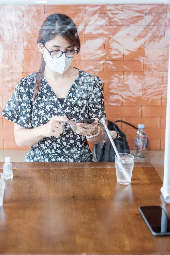 流行病新冠毒社会的亚洲妇女在餐厅饮期间使用智能手机将防护罩塑料隔开保护共生19年大流行后在社交失常新的正和生活之后的冠状渗透社会图片